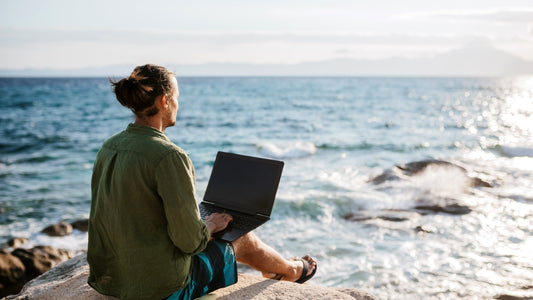 Man with a man bun sitting on a rocky coastal ledge working on a black laptop, wearing an olive green shirt and patterned blue swim trunks, facing a sparkling blue ocean with crashing waves toward the horizon.