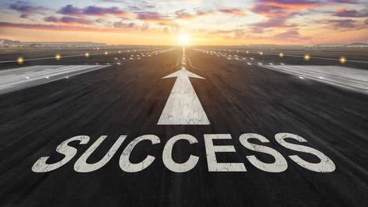 An airport runway stretching toward a vibrant sunset, with the word "SUCCESS" and a white arrow pointing forward painted on the asphalt.