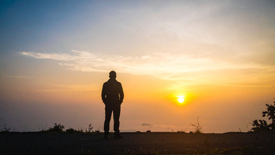 Silhouette of a lone person standing on a hilltop, looking out at a bright sunrise over a sea of mist and low clouds with a warm orange and blue sky.