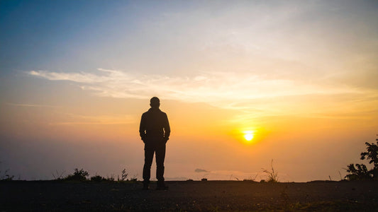 Silhouette of a lone person standing on a hilltop, looking out at a bright sunrise over a sea of mist and low clouds with a warm orange and blue sky.