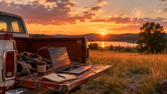 Vintage pickup tailgate with a laptop, coffee, and notebook by a lake at sunset with faint fireworks