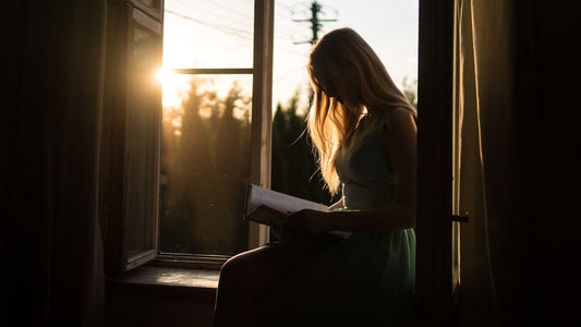 Woman silhouetted by golden window light reading a book alone