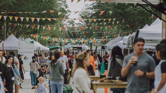 An outdoor festival or market with bunting flags and a crowd browsing white-tent vendors