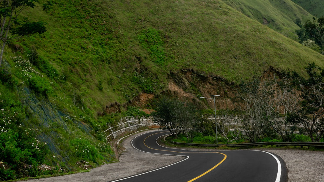 A paved road curving sharply along a green hillside with mountains in the background