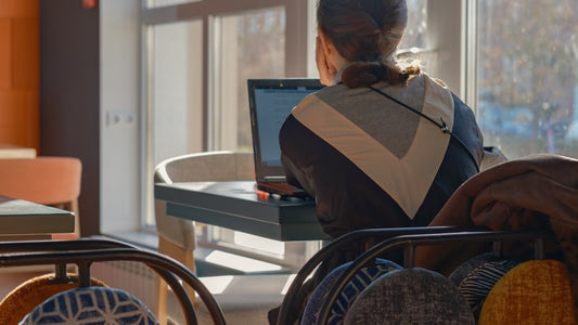A woman working on a laptop at a small table next to a sunlit cafe window