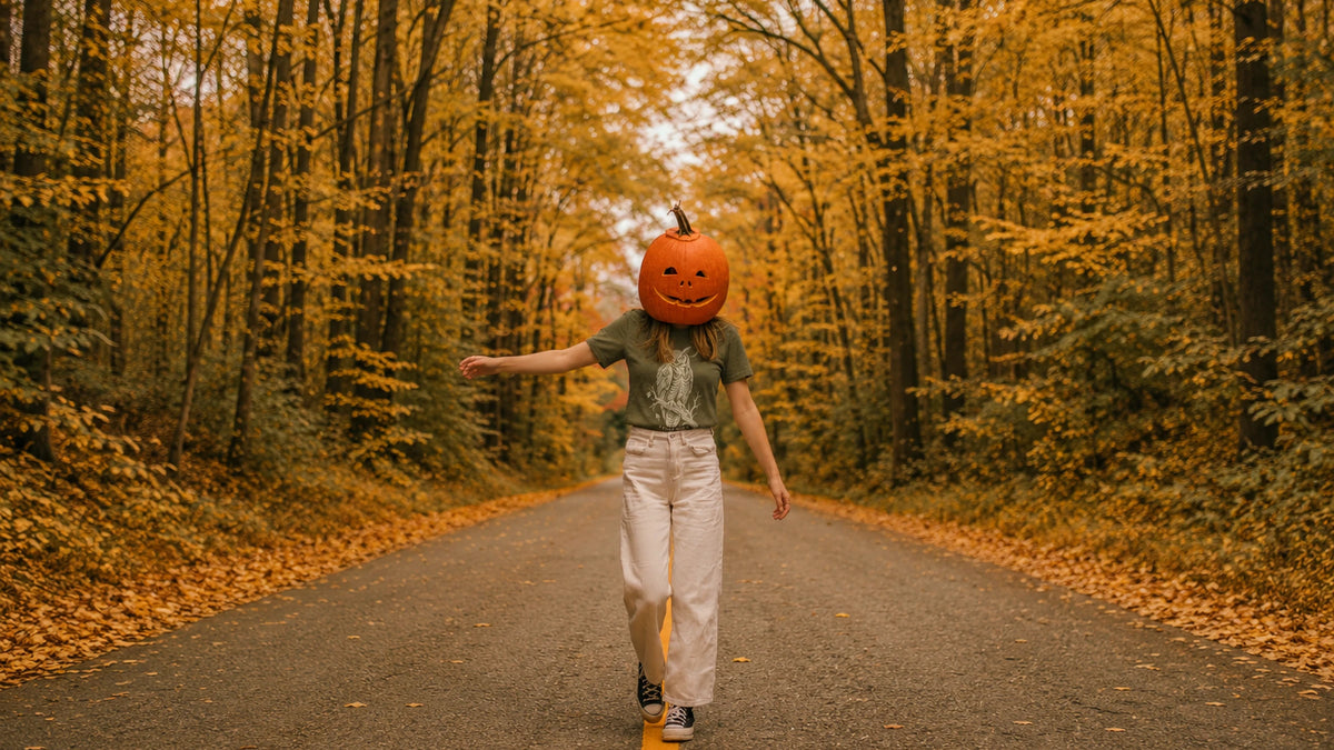 Person with a carved jack-o-lantern head mask walking a golden autumn road wearing the NO.925 Night Shift owl tee