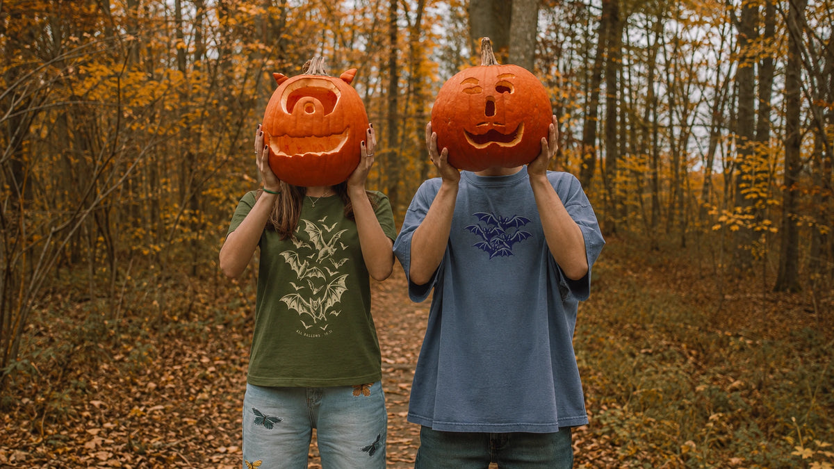 Two people holding carved jack-o-lanterns over their faces on an autumn forest path, wearing NO.925 All Hallows 10.31 and Cluster-O-Bats Oversized Halloween tees